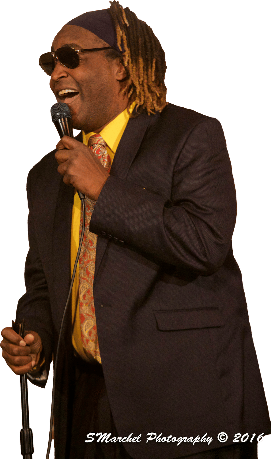 Cool TLC wearing sunglasses, a headband, and a dark suit with a yellow shirt and patterned tie is smiling and holding a microphone.