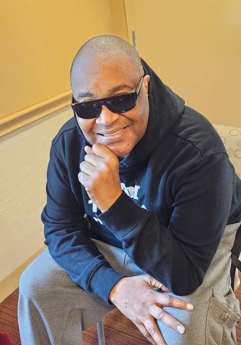Cool TLC wearing dark sunglasses, a black hoodie, and gray sweatpants is sitting on a chair indoors, smiling and resting his chin on his hand.